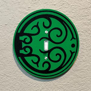 May include: A decorative light switch cover featuring a vibrant green background with a black, swirling design. The cover has a standard toggle switch and is mounted on a textured, neutral-colored wall.