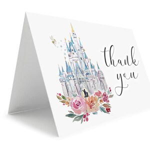 May include: A folded white card with a watercolor illustration of a fairytale castle, flowers, and the words "thank you" in black script. The castle is in shades of blue, gray, and white, with a fairy and stars. Pink and orange flowers adorn the bottom.