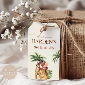 Peut inclure: Une étiquette cadeau avec le texte "Harden's 3rd Birthday" et une illustration de personnages du Roi Lion. L'étiquette est attachée à un cadeau enveloppé de toile de jute avec de la ficelle et ornée de fleurs blanches.