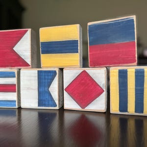 May include: A collection of seven wooden blocks, each painted with a different nautical signal flag design. The blocks feature red, white, blue, and yellow colors. The flags are arranged in a stacked formation, showcasing various geometric patterns.