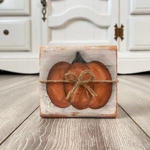 May include: A small, square wooden block with a hand-painted pumpkin design. The pumpkin is orange with a green stem, tied with twine. The block has a distressed, rustic look, perfect for fall decor.