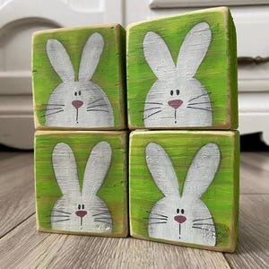 May include: Four wooden blocks stacked, each painted with a white bunny face on a green background. The bunnies have pink noses and whiskers. The blocks are arranged on a wooden floor.