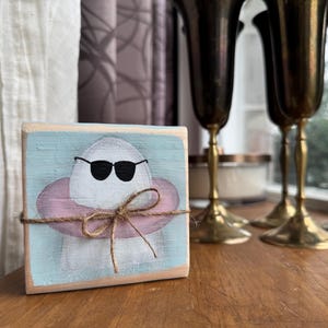May include: A small, square wooden block with a hand-painted design. The artwork features a white egg-shaped figure wearing black sunglasses and a pink inner tube, tied with twine. The background is light blue.