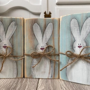 May include: Three wooden blocks with hand-painted white bunny rabbits. Each bunny has a pink nose and is tied with twine. The background is a light blue and brown color, giving a rustic feel. The blocks are arranged on a wooden surface.