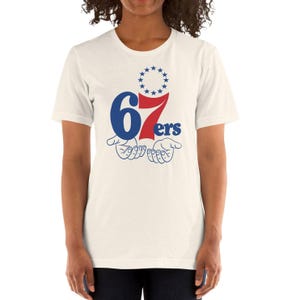 May include: A cream-coloured short-sleeve t-shirt featuring a graphic design with the blue number 6, a red number 7, and the blue text ers. Above the numbers is a circle of thirteen blue stars, and below the text are two open hands.