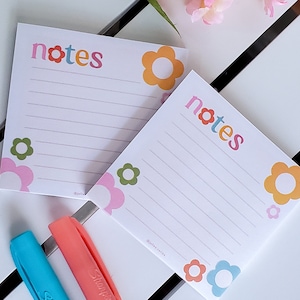 May include: Two pink and white notepads with the word "notes" in colorful letters. The notepads have a floral design with pink, orange, blue, and green flowers.