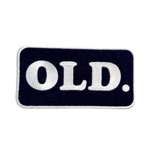 May include: A rectangular embroidered patch with a dark navy blue background and a white border. The word OLD is stitched in large, bold, white capital letters across the center, followed by a white period mark at the end of the text.