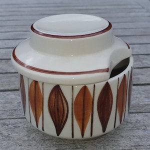 May include: A ceramic butter dish with a lid. The dish is off-white with a brown rim and a pattern of brown and orange leaf shapes. The lid has a small handle. The dish is designed to hold butter and has a small opening for a butter knife.