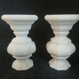 May include: Two white ceramic urns with a decorative, octagonal base and a fluted design. The urns are empty and ready to be filled with flowers or other decorative items.