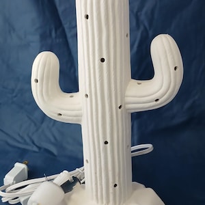 May include: A white ceramic cactus-shaped lamp with multiple holes for light to shine through. The lamp has two arms and a textured surface. Includes a light bulb, a green star, and colorful light bulb covers.