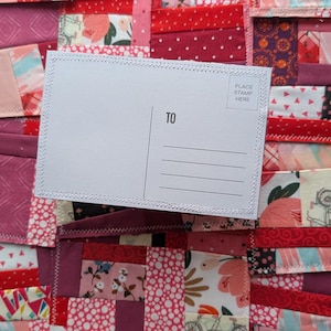 Op de afbeelding: Een witte ansichtkaart met het woord "TO" en lijnen om op te schrijven, geplaatst op een patchwork quilt. De quilt heeft verschillende rode en roze stoffen met bloemen- en geometrische patronen. De ansichtkaart heeft een "PLACE STAMP HERE" vak.