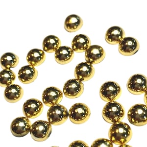 May include: A collection of small, shiny, gold-colored half-spheres scattered on a white background. The objects have a reflective surface, suggesting a metallic or polished material. These decorative elements could be used for crafts or embellishments.