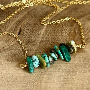 May include: A gold-colored necklace featuring a row of irregularly shaped, vibrant green and turquoise gemstones. The chain is delicate, and the necklace rests on a wooden surface, showcasing the natural colors of the stones.