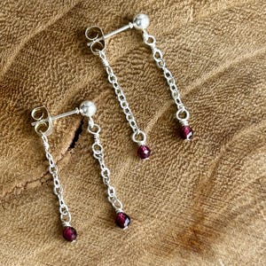 May include: Silver dangle earrings featuring a small stud and a fine chain. Each chain suspends a small, deep red gemstone. The earrings are presented on a textured, light brown wooden surface.