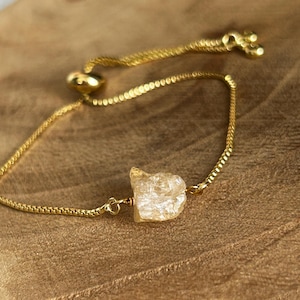 May include: Gold-colored bracelet featuring a raw, light-colored crystal. The adjustable chain bracelet has a delicate design and a sliding bead closure. The crystal is irregularly shaped and adds a natural element to the jewelry.