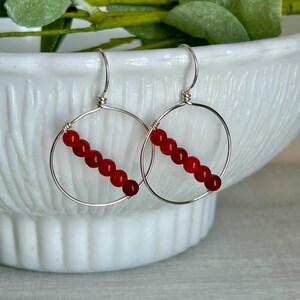 May include: A pair of silver hoop earrings with a row of six red beads. The earrings are displayed against a white background, with a hint of greenery in the upper left corner. The earrings are a simple, elegant design.