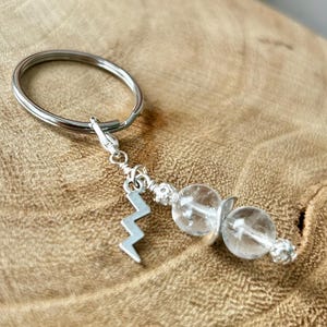 May include: A silver keychain with a lightning bolt charm and two clear, round beads. The keychain is attached to a silver ring. The background is a light brown, wooden surface.