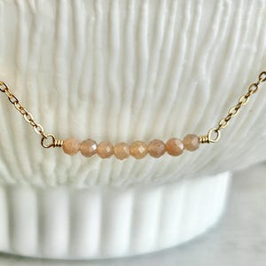 May include: A fine gold chain necklace with a series of small, peach-toned gemstone beads. The beads are consistently spaced and attached to the chain. The necklace is presented against a soft, white backdrop, emphasising its understated elegance.