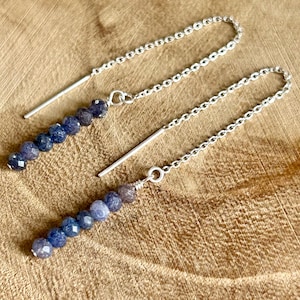 May include: A pair of silver threader earrings, each featuring a delicate chain attached to a straight metal post. At the end of each chain is a vertical row of six small, faceted, deep blue beads that catch the light.