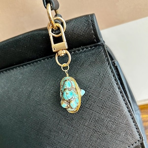 May include: A gold-coloured metal charm with a turquoise stone pendant, attached to a black handbag. The turquoise stone has a unique pattern of blue and brown hues, encased in a gold wire frame. The charm is suspended from a gold clasp.