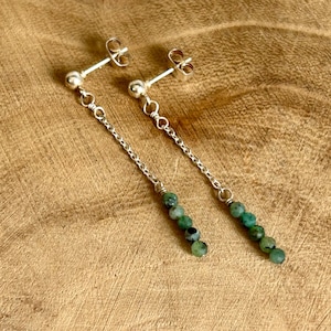 May include: A pair of drop earrings featuring silver-toned posts and thin metal chains. Each earring terminates in a vertical row of four small, round, mottled green beads. The design is symmetrical, with each earring showcasing a consistent length.