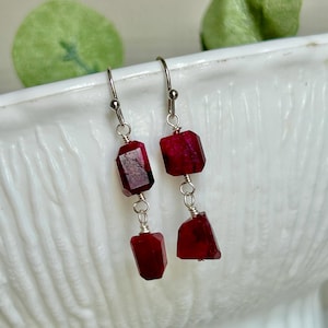 May include: A pair of dangle earrings featuring faceted, deep red gemstones. The gemstones are rectangular and irregularly shaped, connected by silver-colored wire and hooks. The earrings are displayed against a white background.