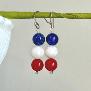 May include: A pair of dangle earrings with a patriotic theme. Each earring features three round beads in red, white, and blue, suspended from a silver hook. The blue beads are at the top, followed by white, and then red.