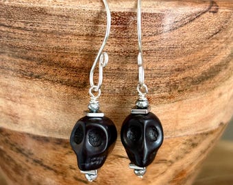 May include: A pair of drop earrings featuring black carved skull beads suspended from silver-toned metal hooks. Each earring includes a small metallic spacer bead above and below the skull, creating a vertical design with a polished finish.