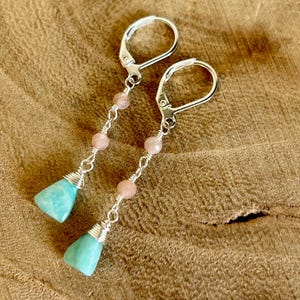May include: A pair of silver leverback earrings. Each earring has a light blue, triangular gemstone wrapped in silver wire, connected to a strand of pink beads. The earrings are displayed on a wooden surface.
