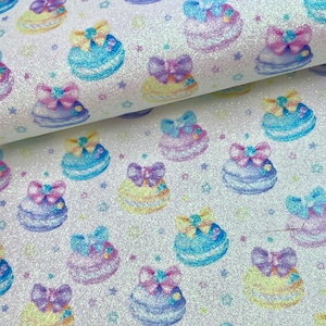 May include: White glitter fabric with a repeating pattern of pastel blue, yellow, and purple macarons with bows. The macarons are scattered on a white background with small white stars.