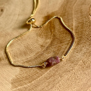 May include: A gold-colored bracelet with a small, rough-cut, pink gemstone. The adjustable chain is delicate and has a small, round clasp. The bracelet is displayed on a wooden surface.