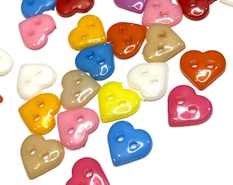 May include: A collection of heart-shaped buttons in various colors, including red, pink, blue, yellow, and orange. Each button has two holes for sewing. The buttons are scattered on a white surface.