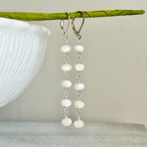 May include: A pair of long drop earrings featuring five small, round, white iridescent beads strung on thin silver-toned wire. Each earring is finished with a silver-toned leverback closure, creating a delicate and linear design.