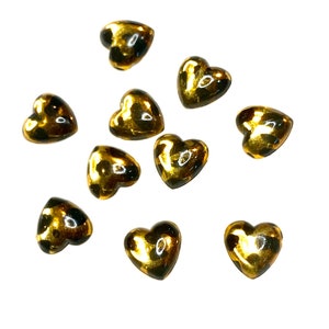 May include: A collection of ten heart-shaped beads. The beads have a glossy, golden finish with dark brown accents. They are scattered against a white background, highlighting their shape and color.