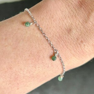 May include: A delicate silver bracelet featuring a chain design with three small, dangling green gemstone beads. The bracelet is shown on a person's wrist, showcasing its simple elegance and subtle color contrast.