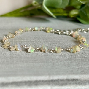 Peut inclure: Un délicat bracelet en chaîne argentée orné de petites pierres d'opale de forme irrégulière. Les opales présentent une gamme de couleurs, dont le blanc, le jaune et des touches de bleu et de vert. Le bracelet est présenté sur une surface gris clair.