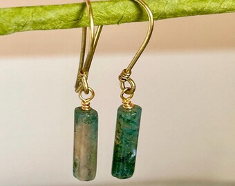 May include: A pair of gold-colored earrings with a hook design. Each earring features a small, cylindrical green and tan stone pendant. The earrings are displayed against a blurred green background.