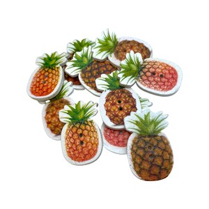 May include: A collection of wooden pineapple-shaped buttons. Each button features a detailed illustration of a pineapple with a green leafy crown and a textured, brown and orange body. The buttons have two holes for sewing.