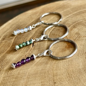 May include: Three keychains with silver-tone rings and clasps. Each keychain features a different beaded charm: one with white beads, one with green and blue beads, and one with purple beads. The keychains are displayed on a wooden surface.