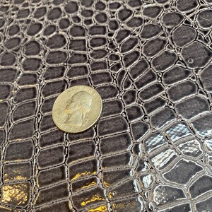 K&ouml;nnte beinhalten: Nahaufnahme eines dunkelgrauen Kunstkrokodillederstoffs mit einer US-amerikanischen 25-Cent-M&uuml;nze oben zum Ma&szlig;stab.