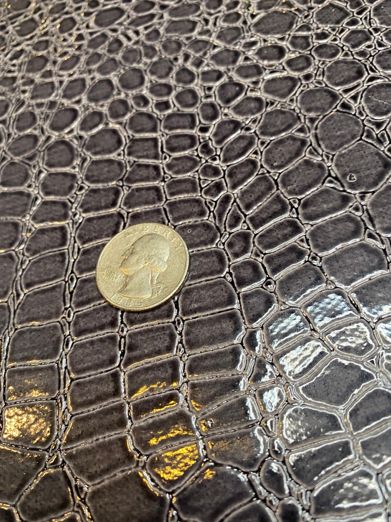 K&ouml;nnte beinhalten: Nahaufnahme eines dunkelgrauen Kunstkrokodillederstoffs mit einer US-amerikanischen 25-Cent-M&uuml;nze oben zum Ma&szlig;stab.