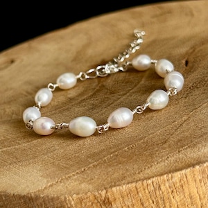 May include: A delicate bracelet featuring oval-shaped white pearls strung on a silver chain. The pearls have a subtle luster and are connected by small silver links. The bracelet is displayed on a wooden surface.