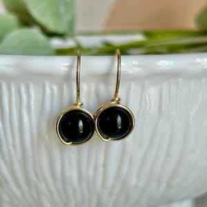 May include: A pair of gold-colored wire-wrapped earrings. Each earring features a round, glossy, black stone. The earrings are suspended from a simple gold-colored hook. The background is a blurred white surface with green foliage.