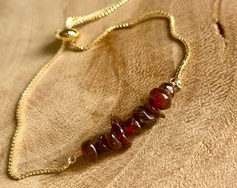 May include: A gold-colored chain bracelet featuring a row of polished, dark red gemstones. The bracelet has an adjustable sliding clasp. The gemstones are a deep, rich red color, and the chain has a delicate, textured appearance.