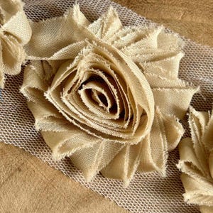 May include: Close-up of a beige fabric flower with frayed edges, layered on a white mesh. The flower is the focal point, with detailed petals and a textured appearance. The background is a light brown surface.