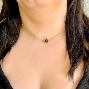 May include: A delicate gold necklace featuring a small, round, deep green gemstone pendant. The necklace is a simple chain design, showcasing the vibrant green of the gemstone. The jewelry is displayed against a skin tone background.
