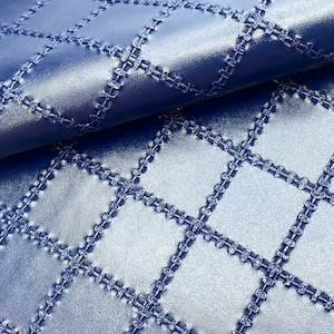 May include: Close-up of a blue quilted fabric with a diamond pattern. The stitching is a darker shade of blue, creating a textured effect. The fabric has a slight sheen, suggesting a leather or faux leather material.