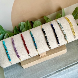 May include: A display of seven beaded bracelets in various colors, including turquoise, purple, red, blue, black, navy, and yellow. Each bracelet features small, round beads strung on a delicate chain with gold-colored accents. The bracelets are arranged on a white, rectangular display.