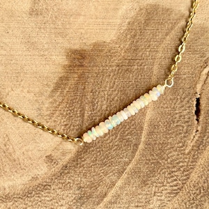 May include: A delicate gold necklace featuring a row of small, iridescent opal beads. The beads are strung together in a linear arrangement, creating a subtle, elegant design. The necklace has a simple chain and clasp.