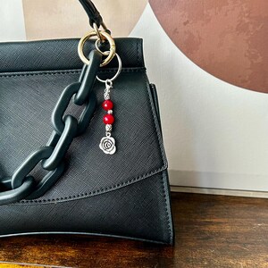 May include: Black handbag with a decorative chain handle and a silver and red beaded charm. The charm features three red beads and a silver rose pendant, attached to a gold-tone ring.